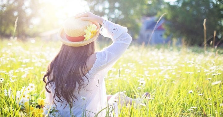 Woman in a straw hat sitting in a sunlit meadow filled with white flowers.